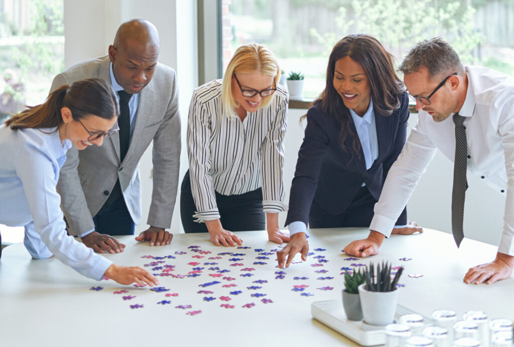 Diverse group of smiling businesspeople standing together in an office trying to solve a jigsaw puzzle on a boardroom table